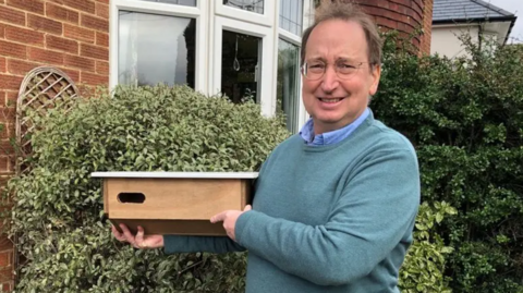 Eddie Bathgate standing in front of a bush outside a home. He is holding a nest box and wearing a green jumper over a blue shirt. He is smiling and looking into the camera. 