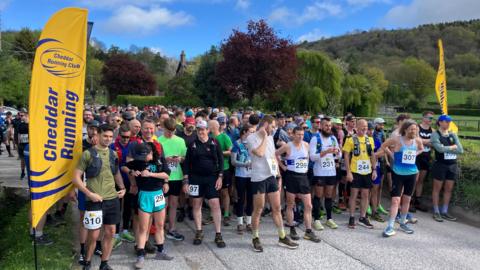 Hundreds of people dressed in running gear lined up at the start line of The Big Cheese race. There are two yellow flags at the start point with the Cheddar Running Club logo.