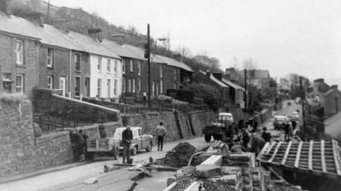 Llun du a gwyn o rhes o dai gyda rwbel yng nghanol y stryd