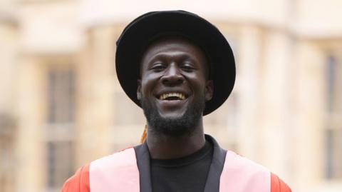 Stormzy is dressed in ceremonial academic attire, featuring a black hat and a vibrant red and pink gown. He is standing and smiling in front of a softly blurred building with pale walls and windows after receiving his honorary degree from Cambridge University.
