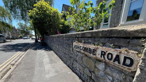 Close up of Ashcombe Road sign which is on a stone wall in front of a house. Road has lots of homes on and green bushes and trees