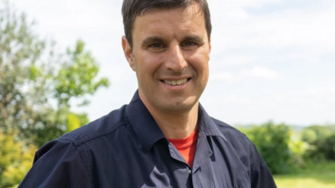 Glynn Luznyj, a man wearing a red t-shirt and navy blue firefighter overalls. He is standing outdoors with fields and trees behind him.
