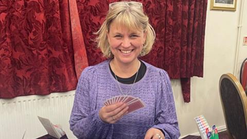 Abbey Smith sitting at a table playing bridge. A pink plastic box is in front of her, with a blue covering on her table. She is holding cars, with other cards in front of her. She is by red patterned drawn curtains. She is wearing a purple top, with a black top underneath, has glasses on her head, and has hair chin length hair. She is smiling. There are two people at the table with her, but you can only see their hands. Another person is sitting down, with their back to the camera to the right. 