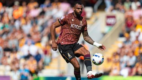 Curtis Tilt in action for Bradford City. He is wearing a maroon and gold striped football shirt with black shorts and socks. A stand filled with football supporters is visible in the background.