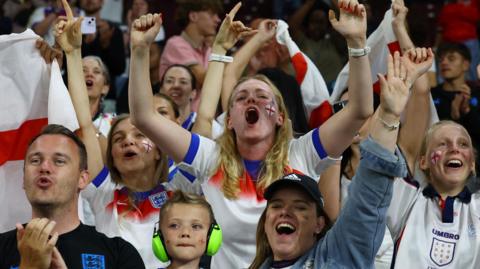A crowd of England fans dressed in England jerseys and wearing red and white face paint celebrate after the match 