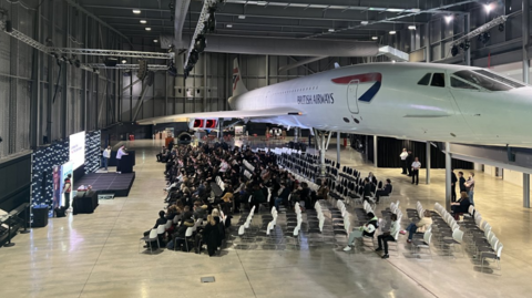 The large hangar at Aerospace Bristol, housing the Concorde aeroplane, with rows of seats and students looking at a temporary stage