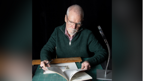 Richard Adams, holding a pencil and sat a desk, looking at a book.
