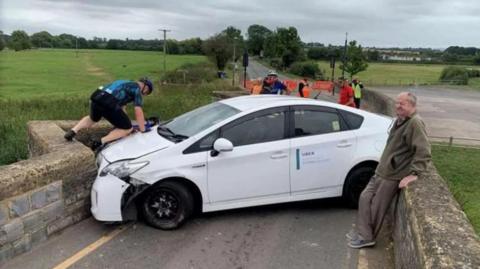 A white Uber car wedged on a 15th Century bridge on the B4085 Honeybourne Road, Bidford-on-Avon. A man in cycling gear is on the car's bonnet, with another man leaning against the bridge, which has red barriers at one end