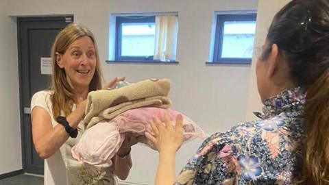 A woman holding a number of items that look like blankets, with a woman handing them to her. They both have long hair.