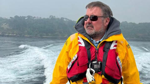 A person standing on the deck of a moving boat, wearing a bright yellow waterproof jacket and a red RNLI-branded lifejacket. The boat is creating a visible wake in the water behind. In the background, there is a coastline with trees and greenery under an overcast sky.