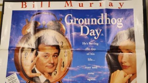 A film poster for Groundhog Day. It features the name of the main actor Bill Murray along with the title of the film. Underneath, it reads: "He's having the day of life life...over and over again." On the left is a cock with Billy Murray's face in the middle. On the left is the actress Andie MacDowell.


