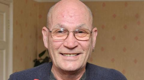 David Newton is smiling at the camera in an image from 2016. He is bald, has some stubble and wears thin framed glasses. He is wearing a navy jumper, with a grey top underneath and has an earring in his left ear. He is stood inside a room that has been papered with rust colour wallpaper that has red diamond shaped patterns. There is a white door behind him to his left and a green plant. 