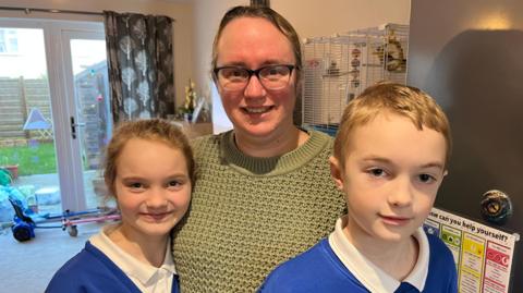 Image shows mum of two, Debbie Brouard, dressed in a dark green jumper, with hair tied back and wearing glasses, hugging her two children, Riva-Mae and Kian, wearing their blue and white school uniform. They're standing in the kitchen, by the fridge, smiling, with bird cage and garden visible in the background.