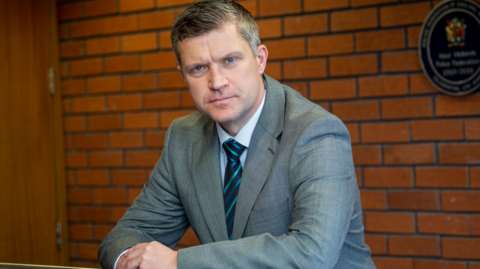 Mr Cooke is wearing a grey suit and is leaning on a bannister with a red brick wall behind him.