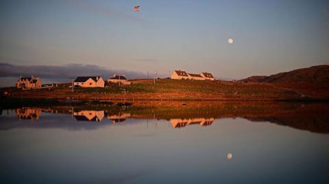 Houses in sunlight and reflected in a loch's waters. There is a full moon and a gull flies above the loch.