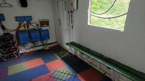 A room with a broken window and a wooden board, which has fallen on a chequered carpet.