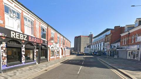Google street view of Victoria Street South in Grimsby town centre. There are shops on either side most in two-storey red brick buildings. In the distance is a white building and a concrete tower block