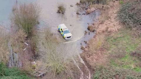 Police 4x4 drives through deep flood water in a field of bushes and other flora. Tyre tracks are visible on the edge of the water and the police vehicle appears to be following them.