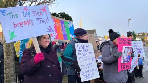 Teachers protesting outside a school, holding signs about cutting pay