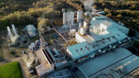 Plumes of steam rise from the cooling towers of a factory complex, seen from above.
