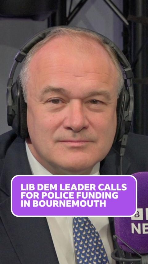 Ed davey wears headphones and looks at the camera, he has balding grey hair and wears a white shirt, blue and white patterned tie and a dark blue blazer.