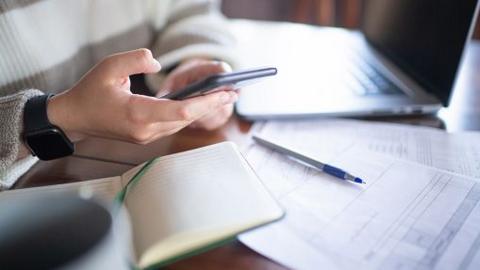 Young woman with smartphone and laptop reviewing banking or financial papers