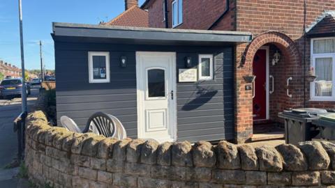 The shed has a door and has a flat roof. It is painted black and sits next to the front door of the house. 