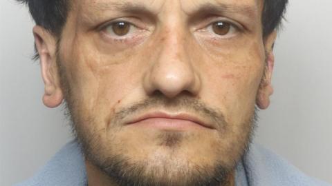 A man with a dark beard stares into the camera in a police custody shot.