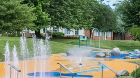 A water play area in a park. There is a yellow and blue floor with water fountains dotted across it. There are lawns and trees in the background, as well as some houses that are partly visible on the edge of the park.