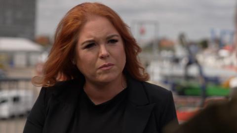 Hannah Damary-Wilson. She has ginger hair and is wearing a black jacket and black top. 
