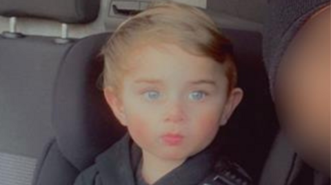 A child in a car with a seatbelt on. He is wearing a black hooded top with "Nike" written on the front. 