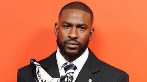 Skepta at the GQ Men of the Year awards. He is wearing a black suit and stood against a bright orange background.