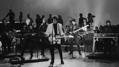 Tandy is on stage with other band members in this black and white photo. He is sat at a keyboard while other band members sing and play instruments.