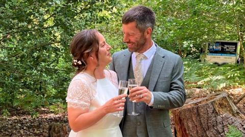 A man, dressed in a suit, and a woman in a wedding dress toast champagne in a woodland.