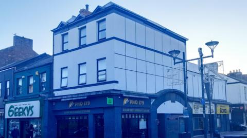Photo of two-storey building above a restaurant which says "Pho 179". Next to the restaurant is a place called "Geekys". The two-storey building is white and has three windows on each floor. The walls are white. There is street lighting in front of it.
