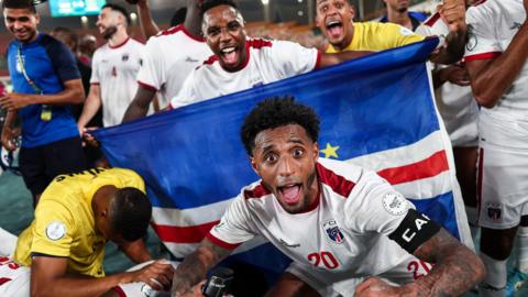 Cape Verde footballers celebrate a victory, with captain Ryan Mendes, wearing a white shirt with red trim, crouching down and extending his arms in front of a national flag and pulling a wide-eyed face of celebration with his mouth wide open