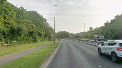 A dual carriageway curves away, with a separate footpath alongside the road. Grass between the road and the footpath. Tall hedges on either side of the road.