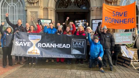 A group of men and women stand outside a council building, cheering, holding banners that read 'Save Roanhead'.