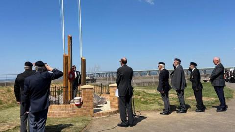 Men in suits and uniform are standing around three pillars that make up a new memorial. They are saluting it.