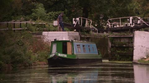 A narrowboat on a canal approaches a lock, while a person stands beside the lock gates. Trees and vegetation line the waterway in the background.