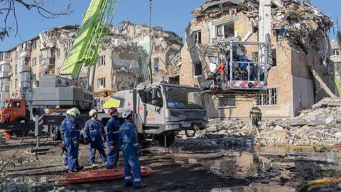 Emergency workers attend to damaged buildings - they're wearing blue overalls and silver helmets