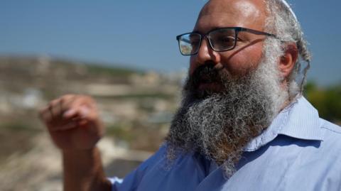 Yehuda Shimon, a large, bearded man wearing glasses, gestures with his hand