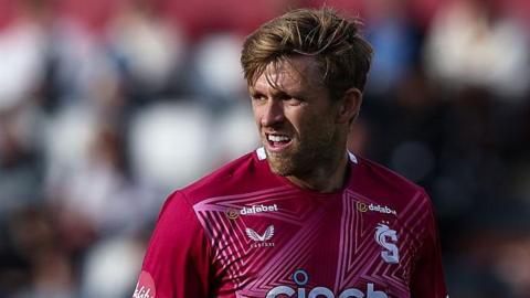 David Willey in action for Northamptonshire in a T20 game. He is looking to his right and wearing a red and pink shirt