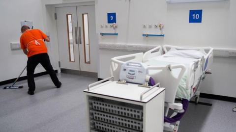 A man with his back to the camera can be seen sweeping the floor in what looks like a clean hospital room, featuring a bed with sides on it. The room also has a variety of plug points on the wall and a medical-looking cabinet at the foot of the bed.