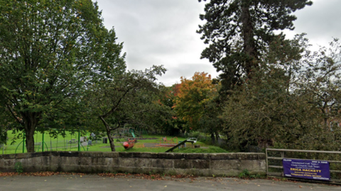 Small play park in the distance with slide and swings, a river on the right of the park and trees in the background.