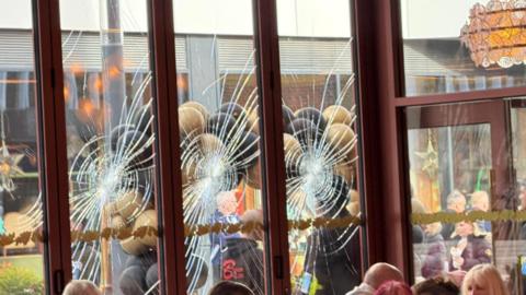 A close-up of a few windows that have been smashed. There are gold and black balloons visible, and various groups of people.