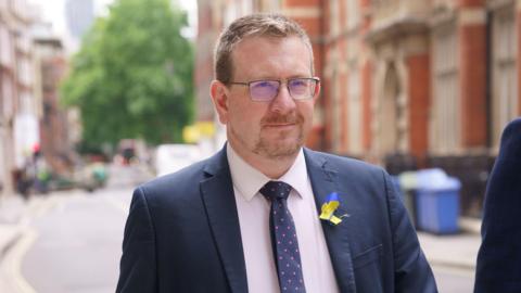 Andrew Gwynne walking a street wearing a suit and a Ukraine ribbon on his lapel. 