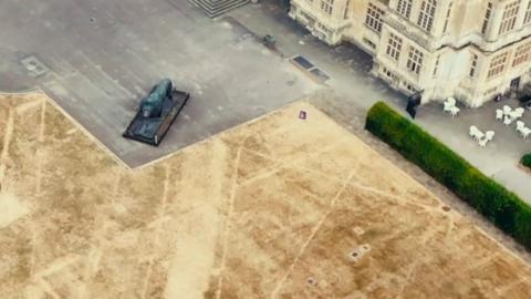 An aerial shot of a parched area of dry yellowing grass in front of a stately home