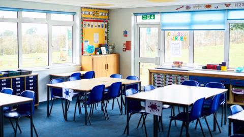 An empty classroom at a primary school in Godalming in Surrey. Blue chairs are placed around various tables. In the background are windows, through which grass can be seen. There are also cupboards, including space for trays.