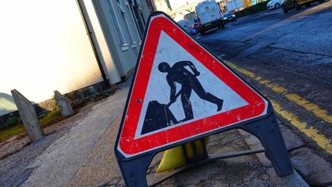 A triangular roadworks sign with a red border by the side of a residential road 